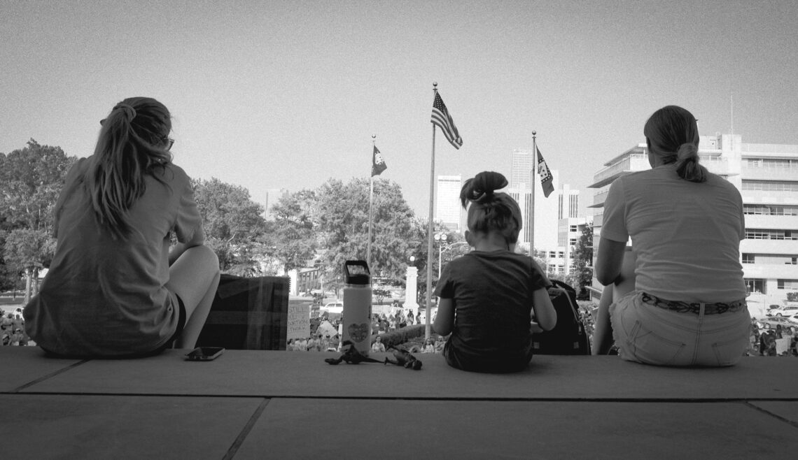 Black and white photo of the backs of three generations of women sitting on top steps of the Arkansas state Capitol under the American flag