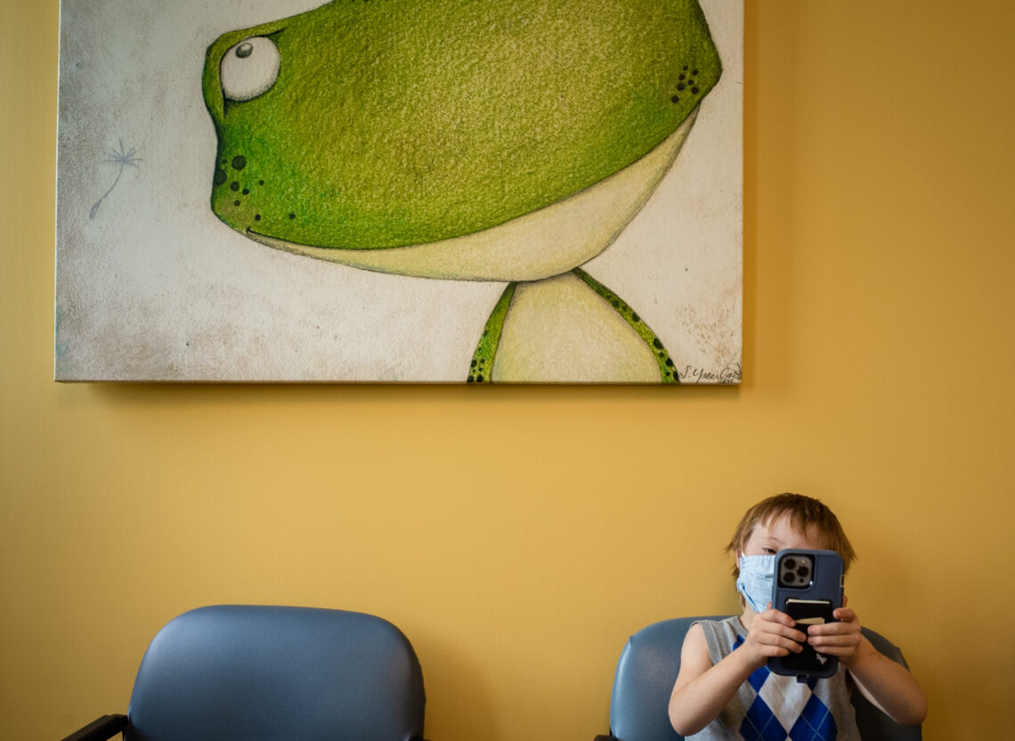 color photograph of boy in blue chair against yellow wall under green frog taking a video of photographer in a doctor's waiting room in Little Rock, Arkansas, that is part of the story we tell project.