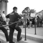 Black and white street documentary photograph of two little rock arkansas police officers standing on the steps of the Arkansas Capitol looking at a black man in front of a crowd of protesting women and their guns at the ready during the rally for women's reproductive autonomy in Little Rock, Arkansas on June 24, 2022